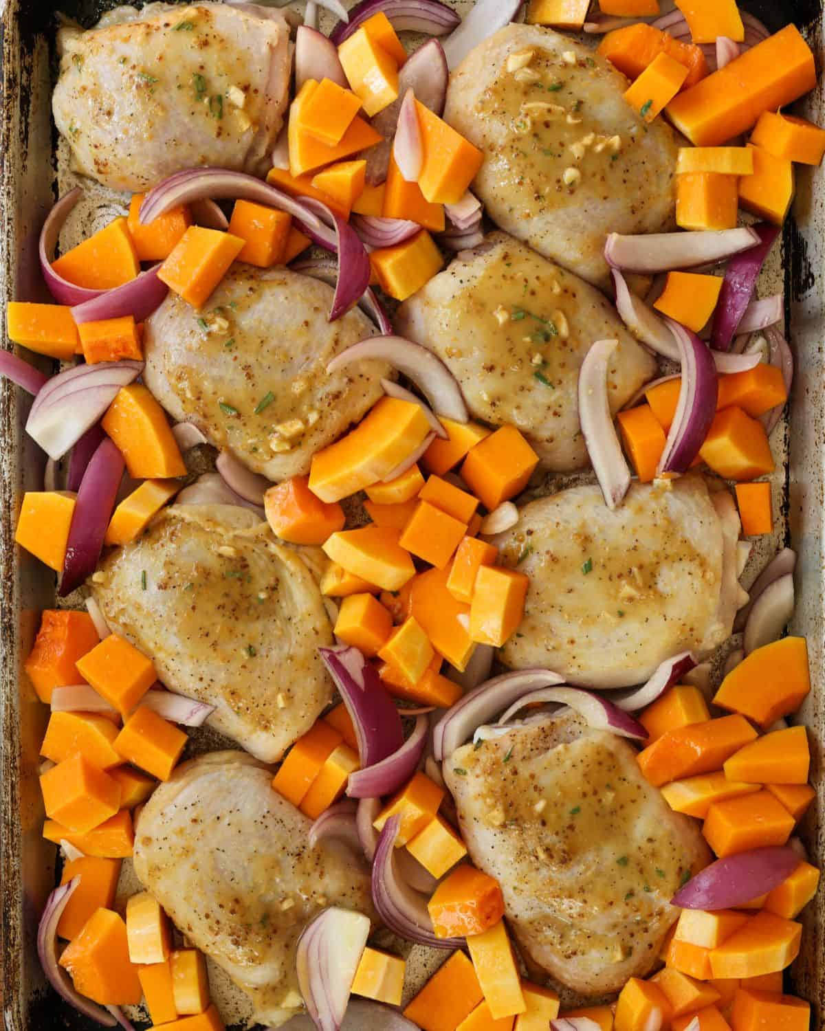 Partially cooked chicken thighs with butternut squash and onions on a baking sheet.