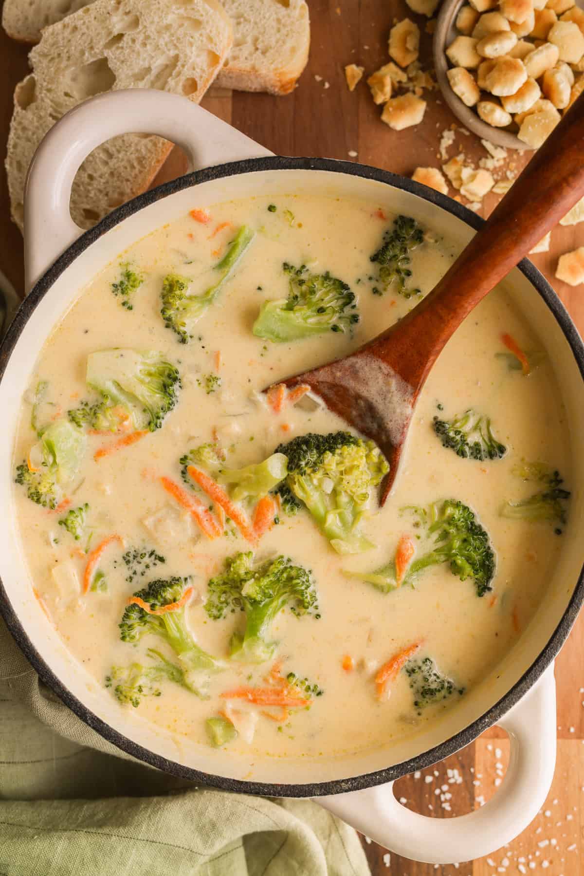 A Dutch oven with creamy chicken broccoli cheddar soup with a wooden spoon. Soup crackers and fresh bread are on the side.