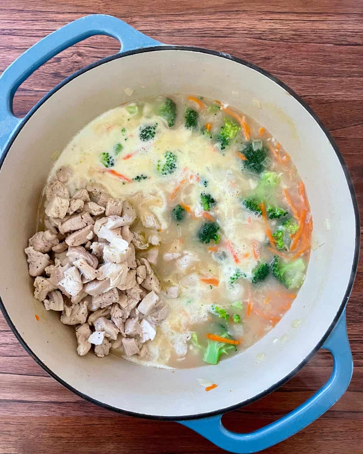 Chicken broth, heavy cream, broccoli, carrots, and cooked chicken are added to the onions, all cooking in a Dutch oven.