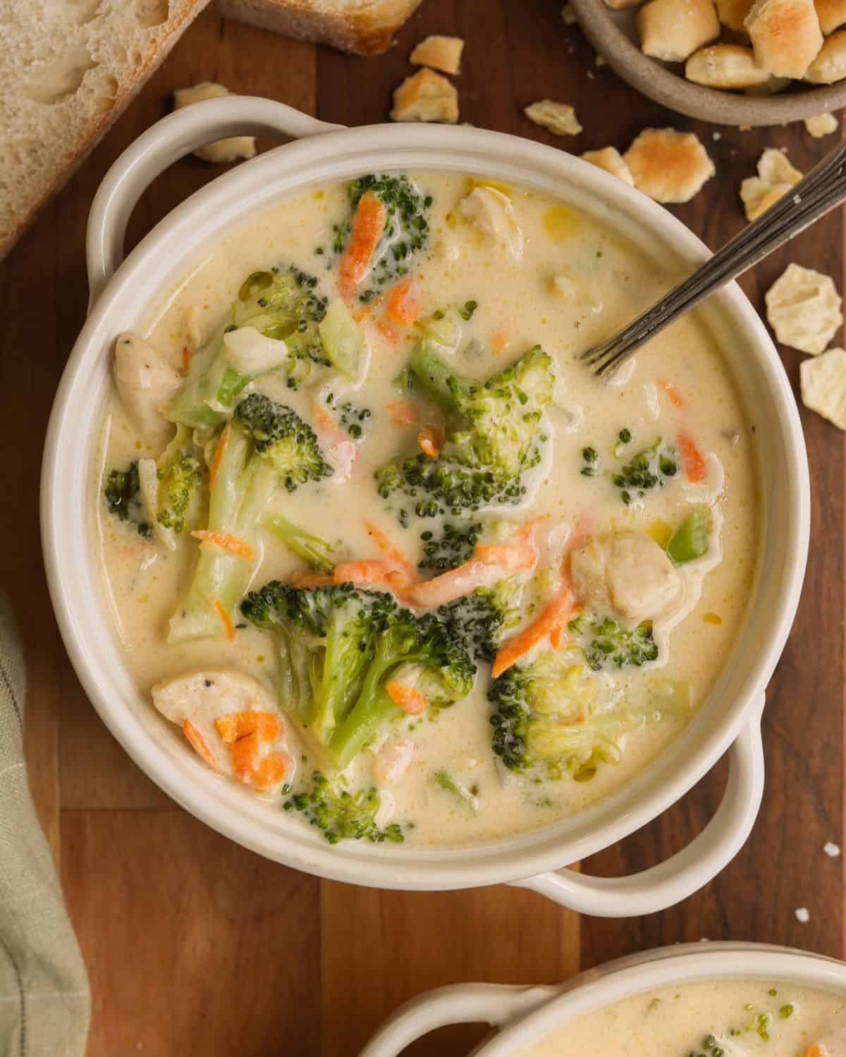 A small bowl with a spoon of broccoli cheddar soup with chicken breast. Crackers and bread are on the side.