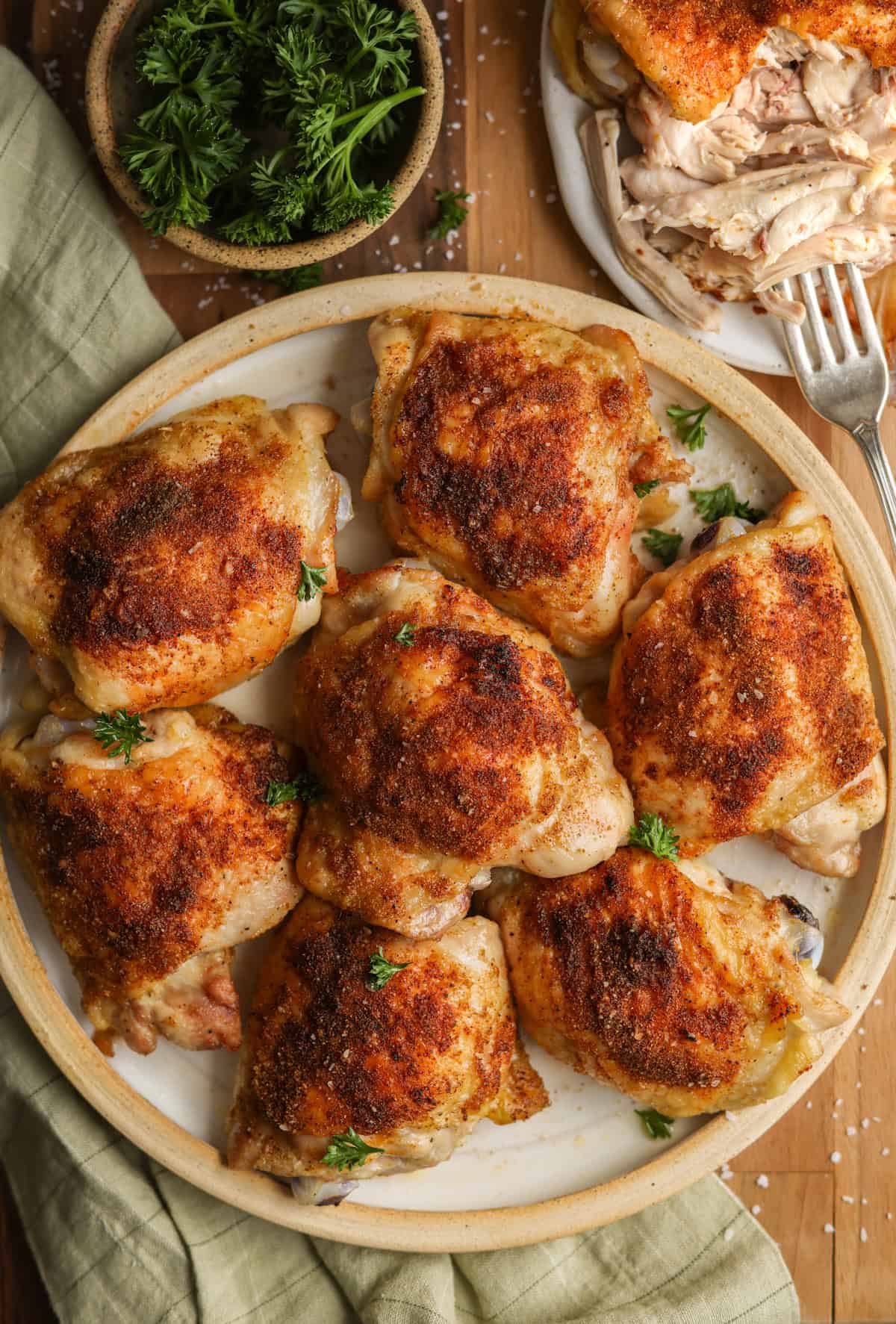 Oven baked chicken thighs on a plate, ready to serve. A small dish of parsley for garnish is in the corner, along with a cut up chicken thigh.