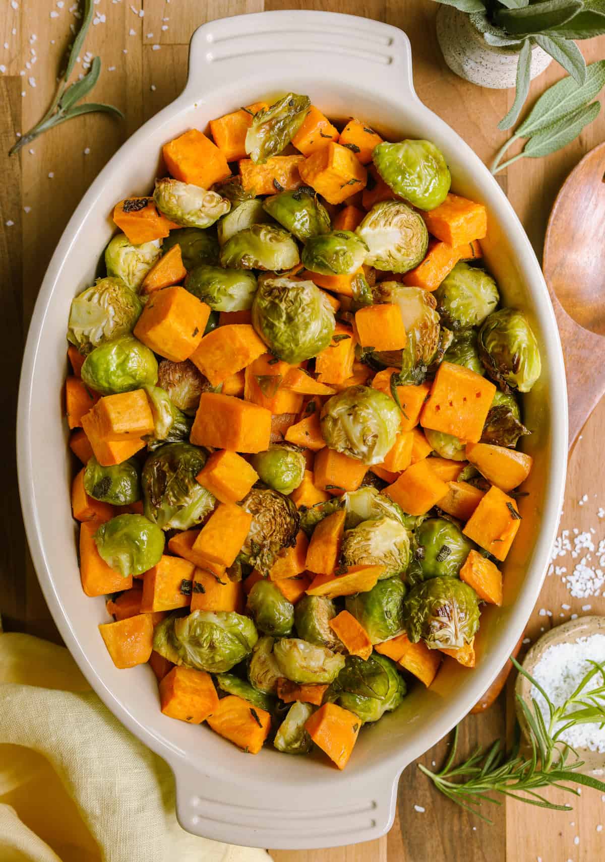 Roasted sweet potatoes and brussel sprouts in a oval-shaped casserole dish, garnished with fresh rosemary and sage.