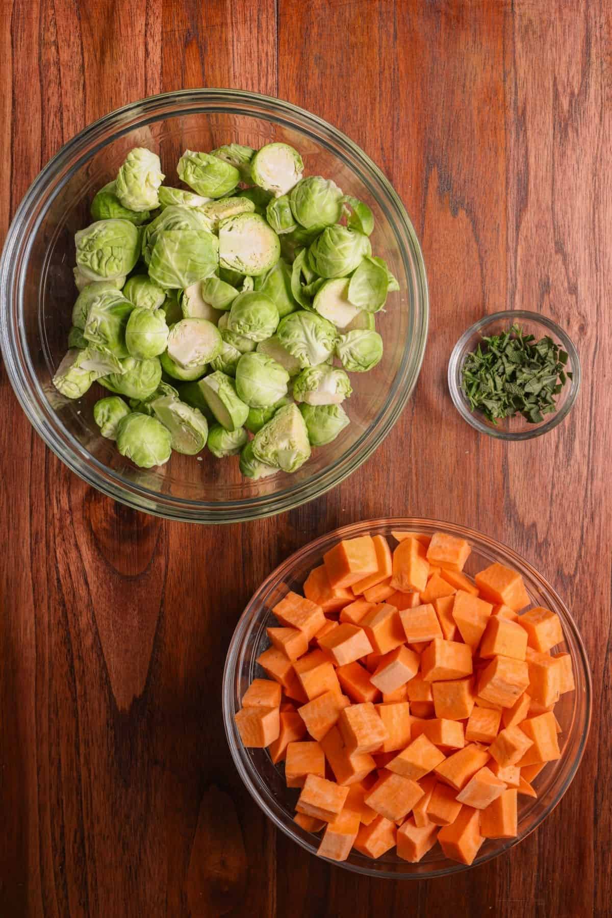 Chopped brussels sprouts in a bowl, minced fresh herbs in a small dish, and peels and chopped sweet potatoes in large bowl.