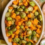 Roasted sweet potatoes and brussel sprouts in a oval-shaped casserole dish, garnished with fresh rosemary and sag.