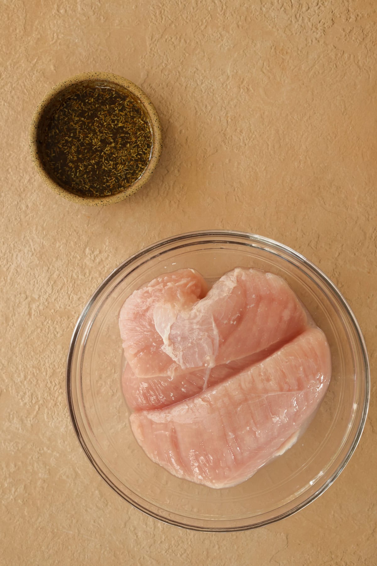 Raw turkey tenderloins in a bowl with a small dish of seasonings mixed with olive oil in a separate bowl.