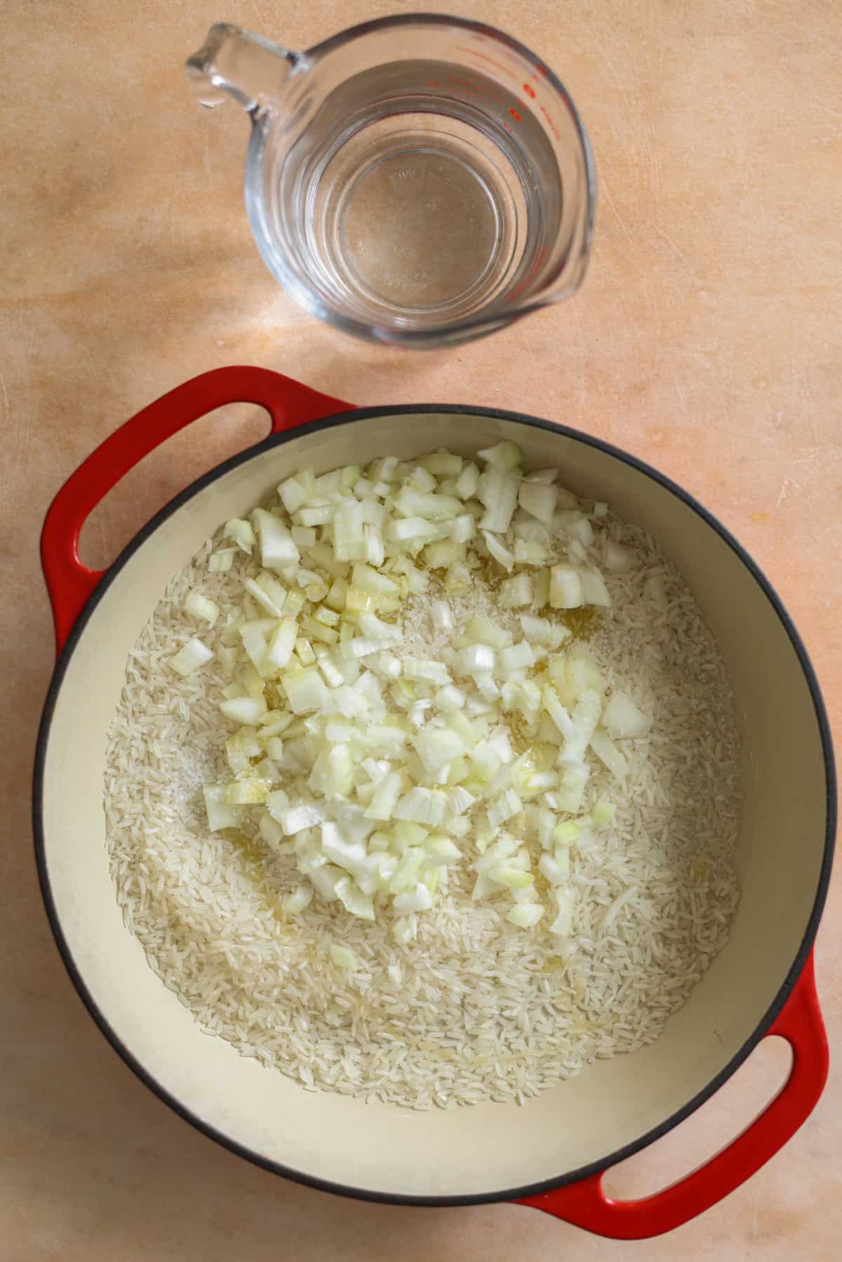 Uncooked basmati rice and diced onions in a red skillet. A measuring cup of water is off to the side.