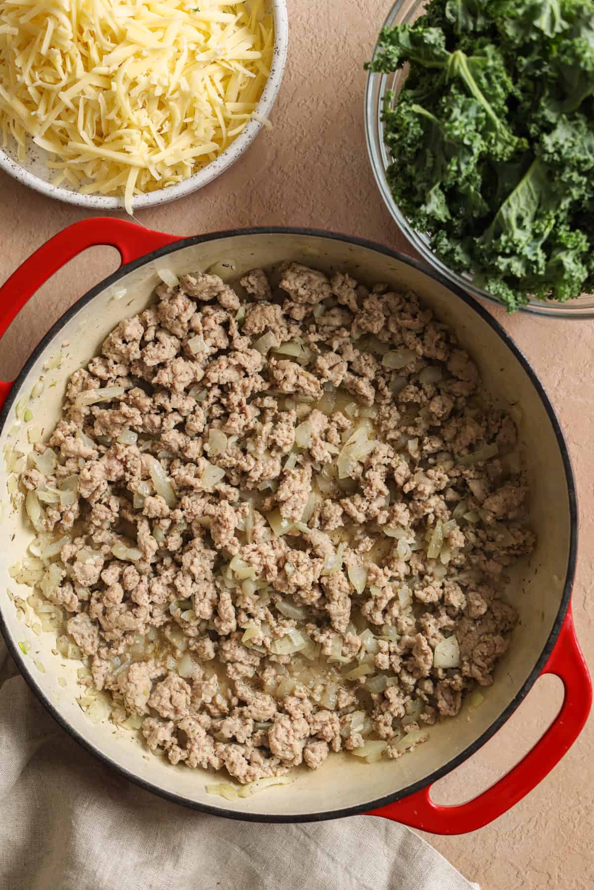 Ground turkey and diced onion cooking in a large pan. There is a bowl of shredded cheese off to the side and a dish of washed and chopped kale.