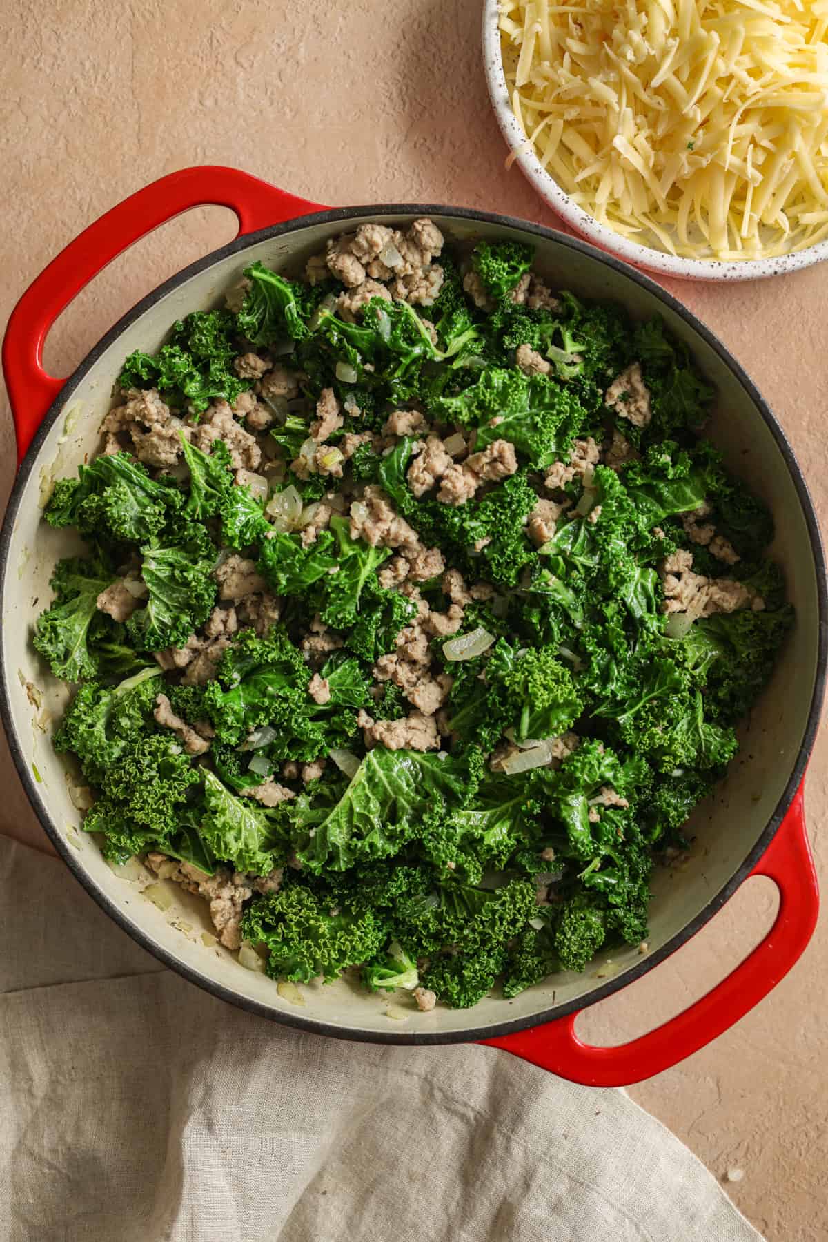 Kale and ground turkey cooked in a red, cast iron skillet, with a bowl of shredded Gruyere cheese on the side.