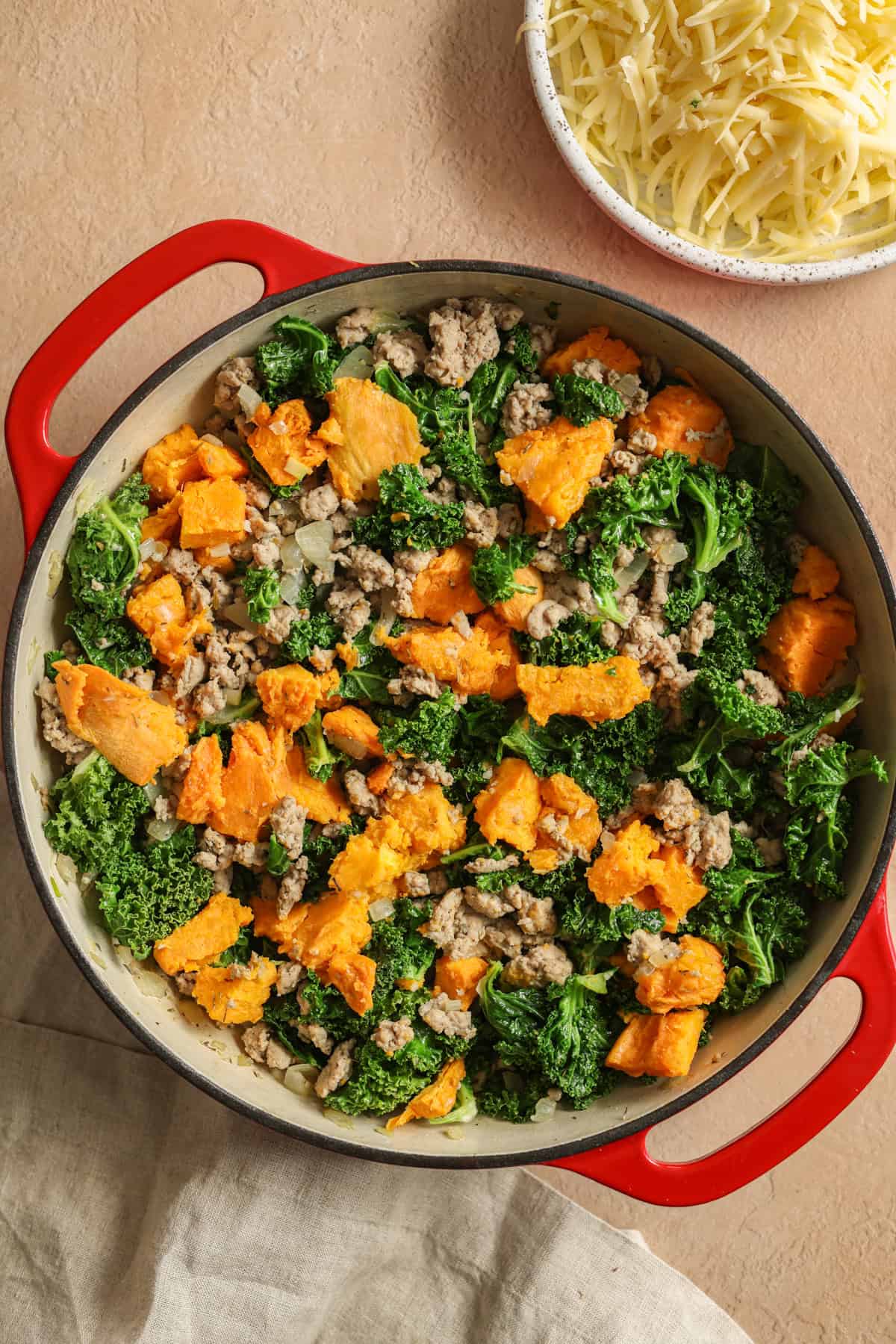 Ground turkey, chopped sweet potatoes, and kale in a large red skillet with a bowl of shredded cheese on the side.
