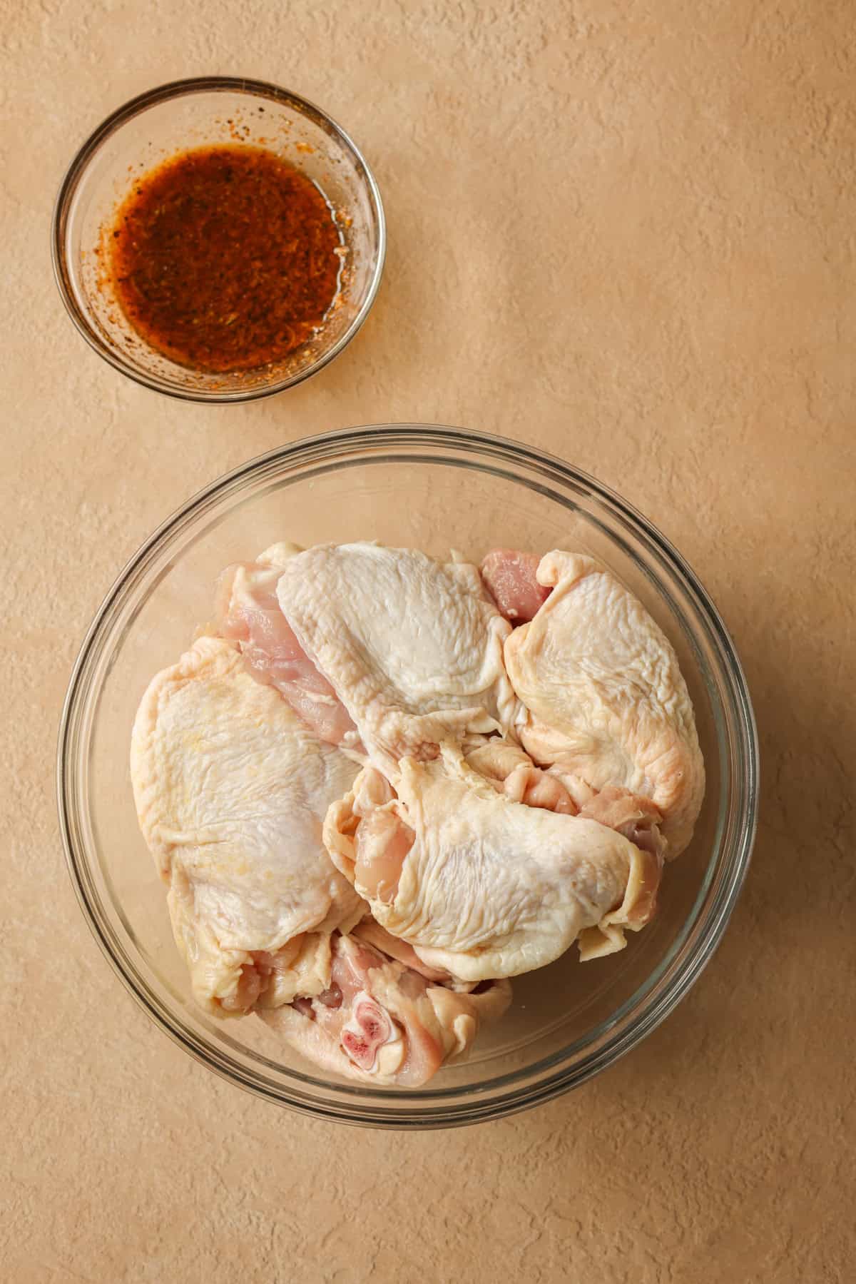 Raw bone-in chicken thighs in a glass mixing bowl with a small dish of lemon pepper rub next to it.