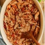 Shredded Mexican-style chicken cooked in a slow cooker. It's in a round serving dish with a wooden spoon.