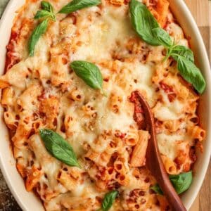 Up close image of a simple baked ziti. There is a wooden spoon scooped into the center of the pasta dish and it's garnished with fresh basil.