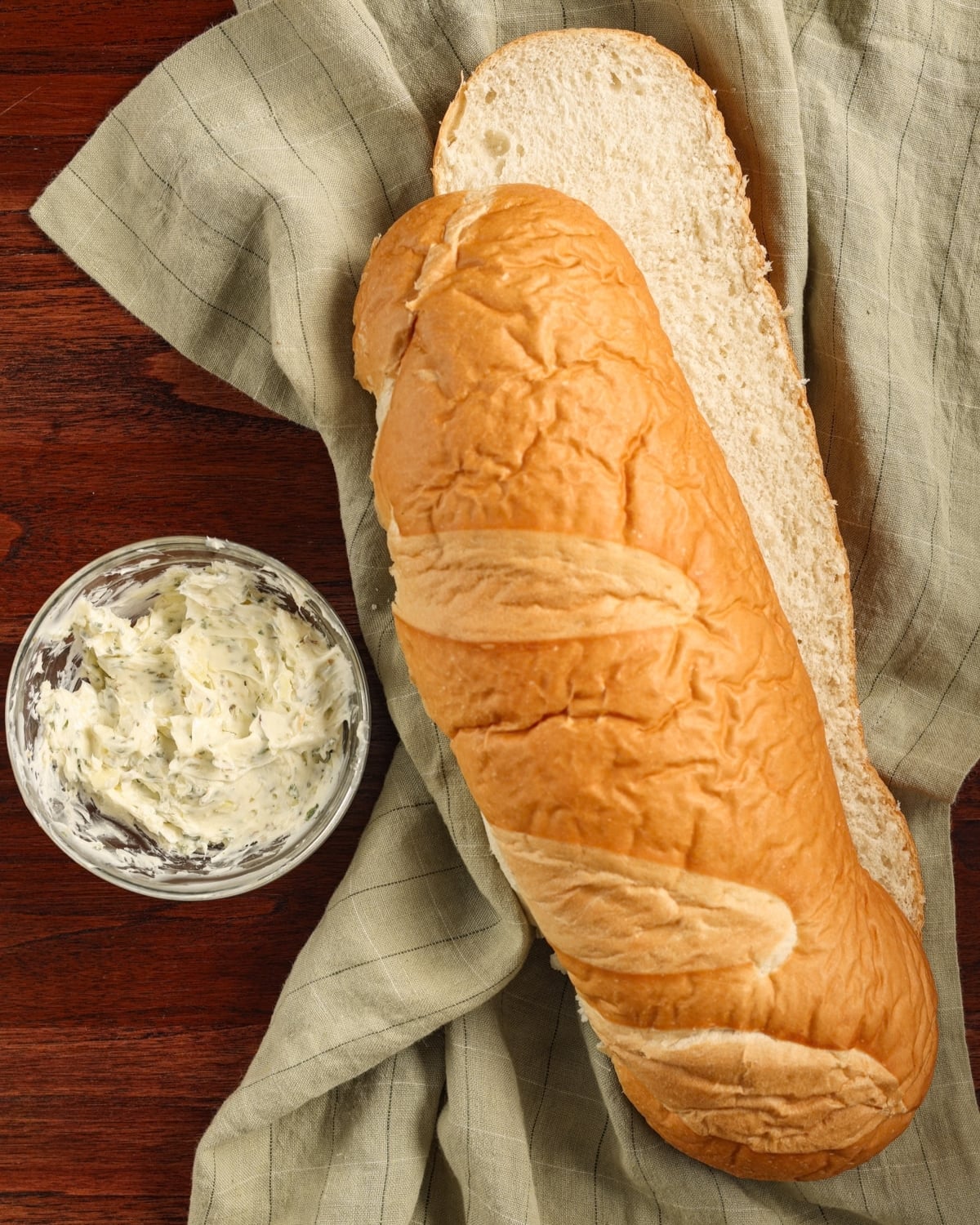 A dish of garlic butter next to a sliced open Italian bread loaf.
